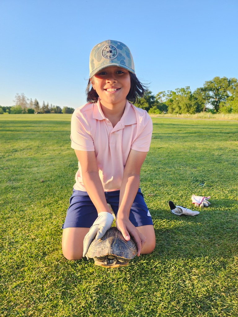 Shay kneeling on a golf course fairway holding a turtle, wearing a golf glove